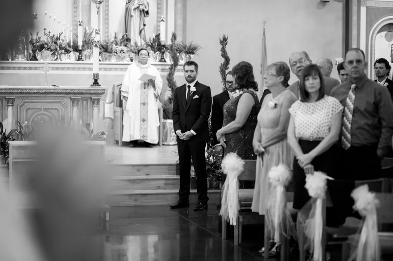Black and white of the groom standing at the altar in his tuxedo while the guests rise from their seats