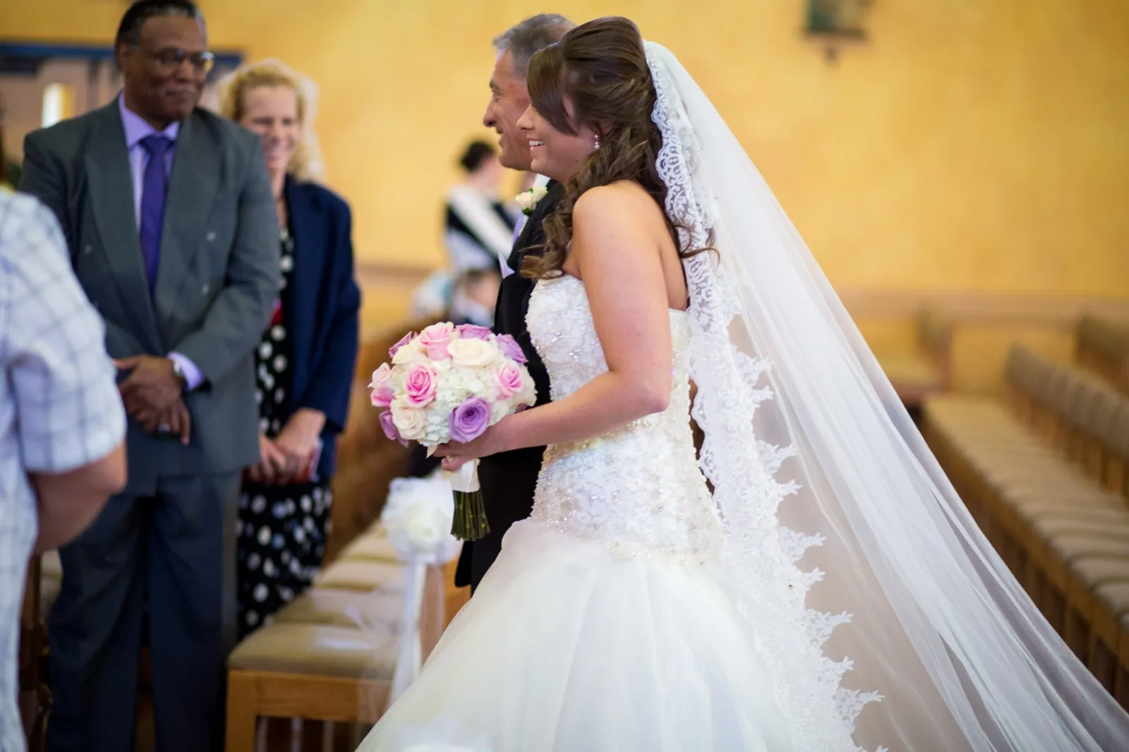 A bride in a long lace edged veil walks up the church aisle on an older man’s arm as guests smile.