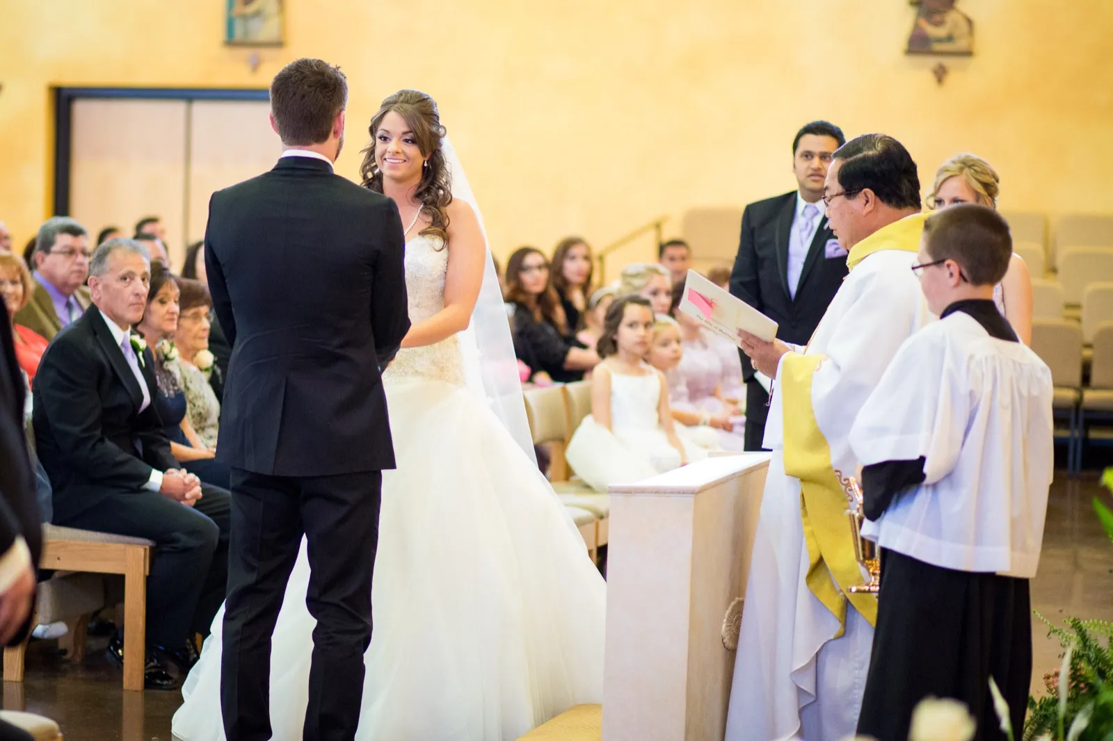 A priest in white and gold vestments reads from a book beside an altar server as a bride and groom face each other