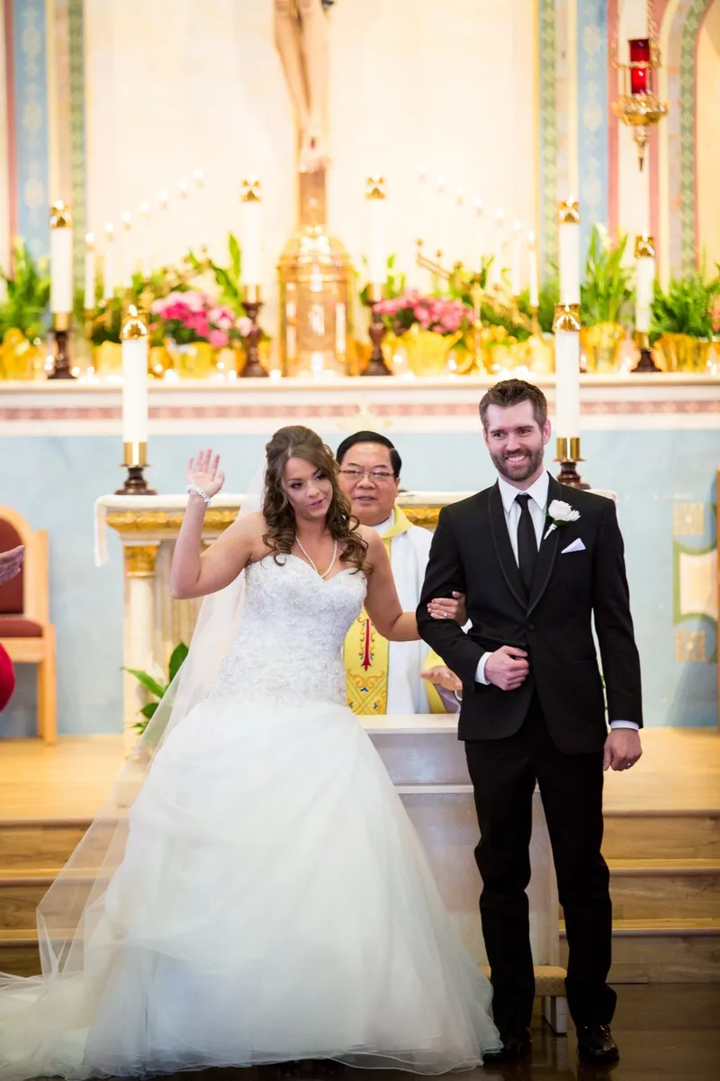 Bride waving to someone in the congregation while she and the groom stand at the altar with the priest between them