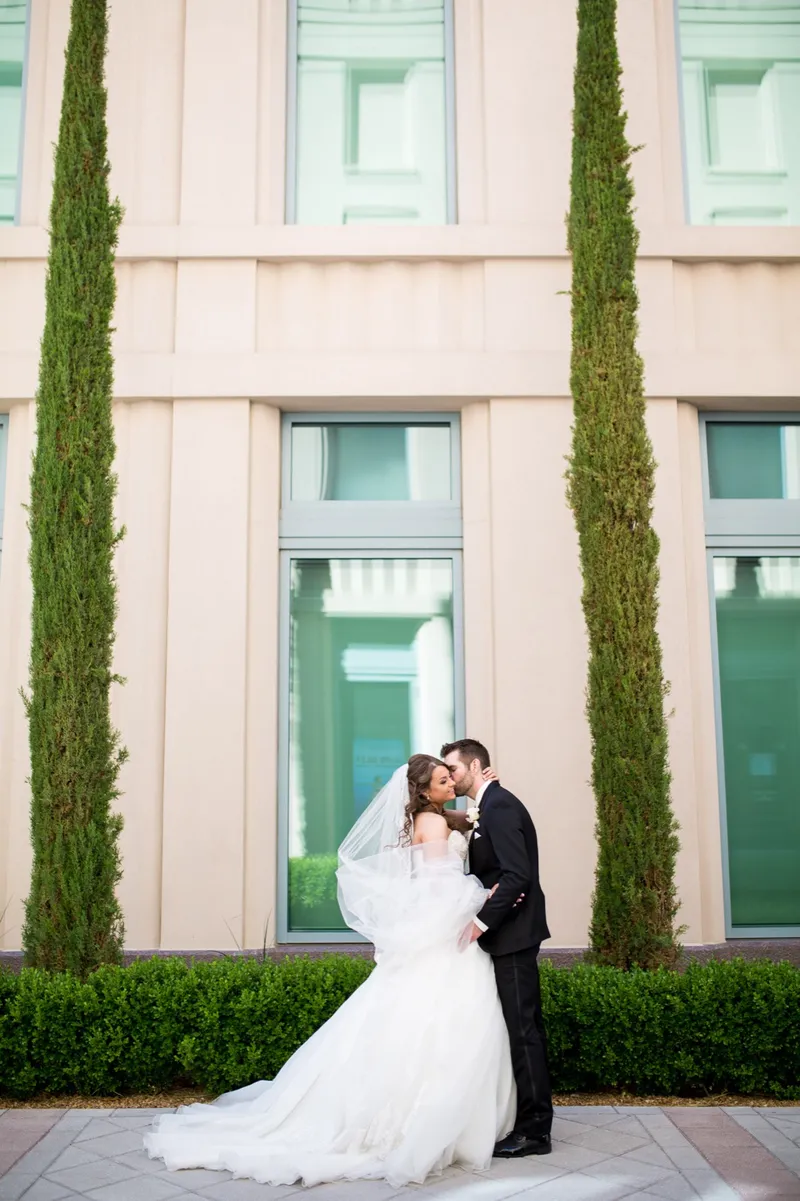 Couple kissing in front of the tall stone facade with a row of narrow cypress on either side of them
