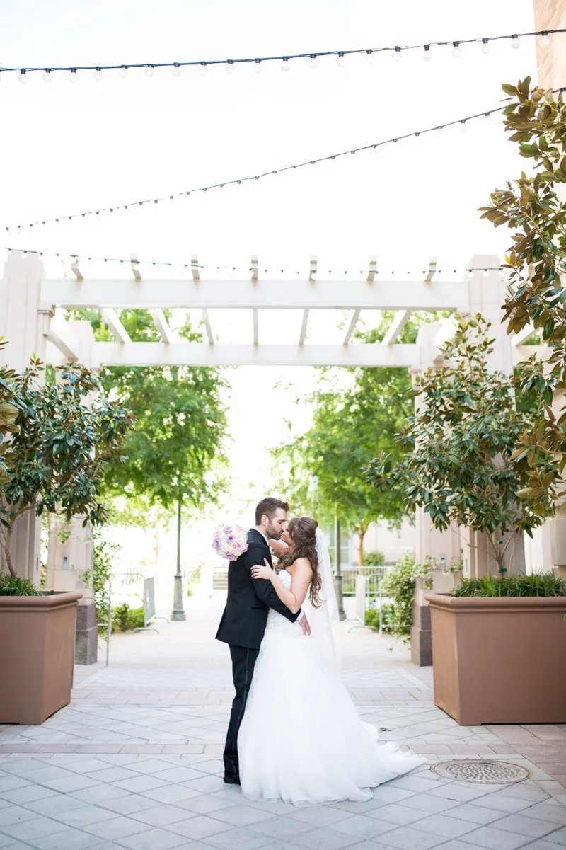 Bride and groom holding each other between the potted trees, cafe lights strung in rows above their heads
