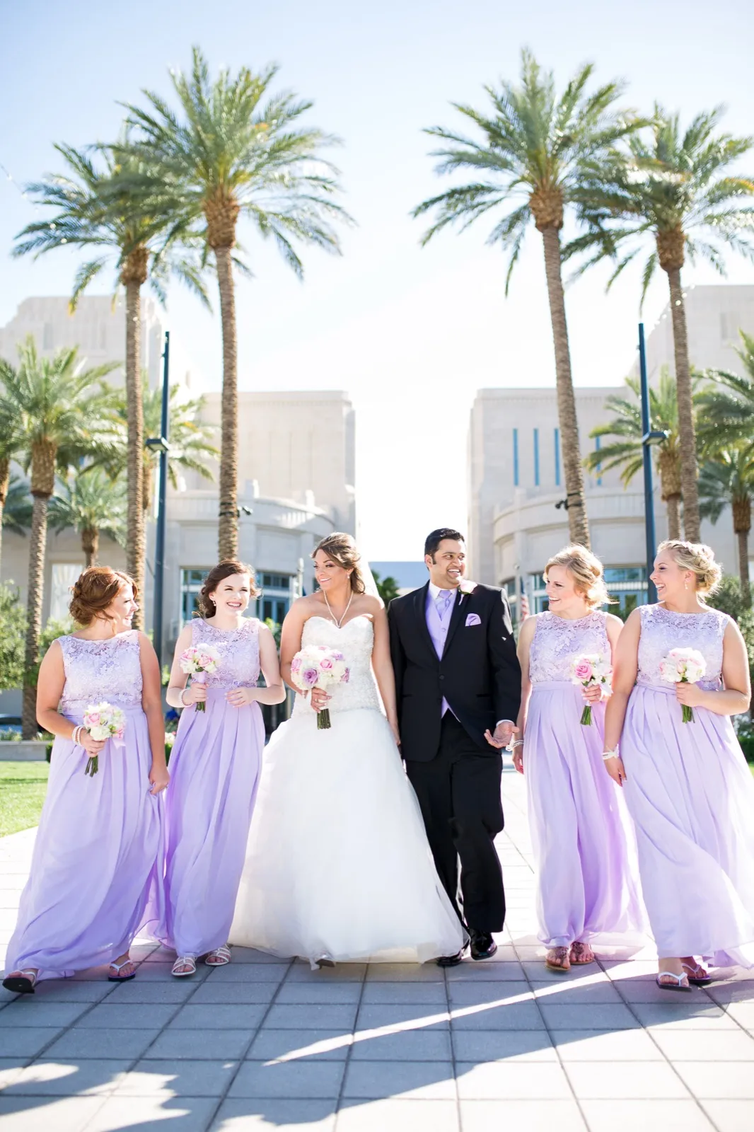 Bride and groom walking a palm lined path with four bridesmaids in lavender gowns spread out beside them
