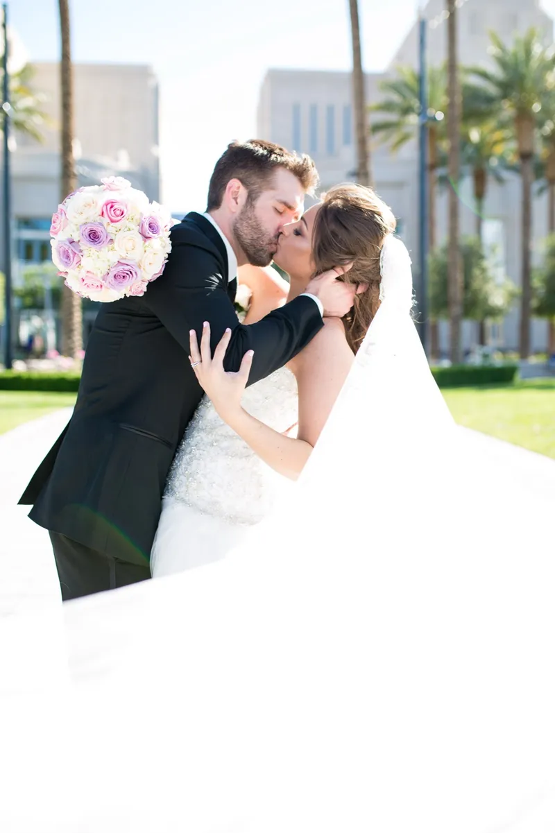 Close frame of the couple kissing with her lavender and white bouquet raised beside their faces, the walkway behind them washed pale in the sun