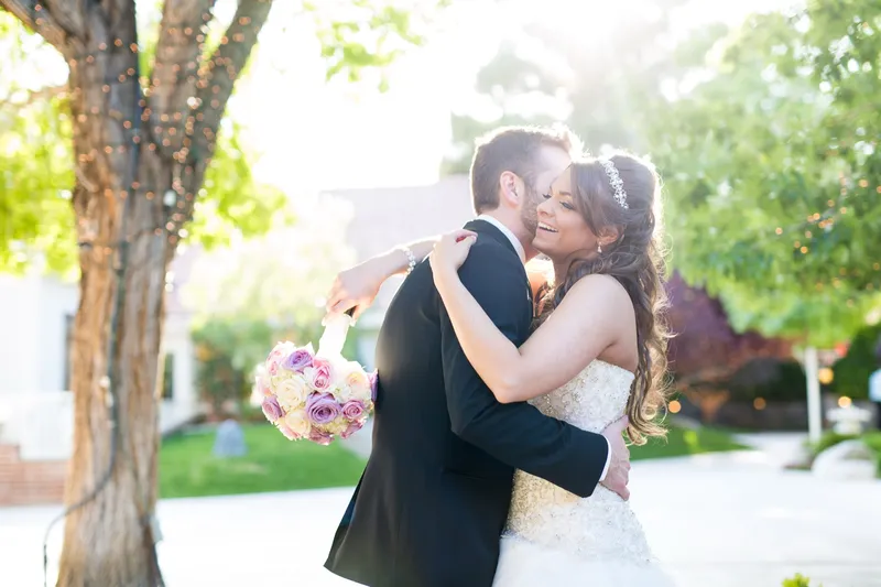 Couple holding each other under the trees in warm late afternoon light, her bouquet raised at his shoulder