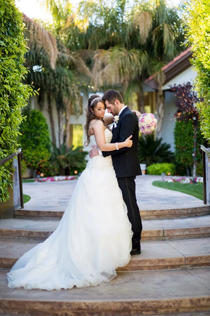 Couple standing close on wide stone steps with tall green hedge walls on both sides and flower beds behind them