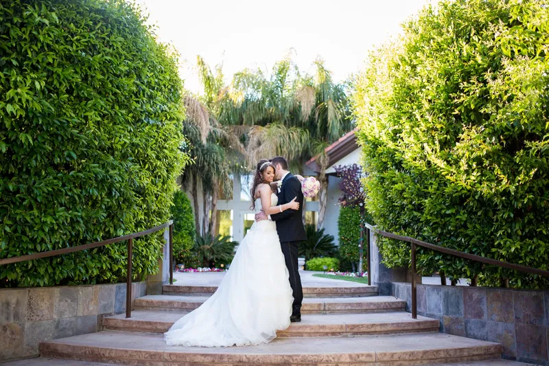 Couple kissing on the hedge lined steps with palms and the venue roofline behind them