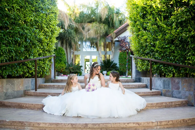 Bride sitting on the wide stone steps with a flower girl on each side of her, tall hedges rising on both sides
