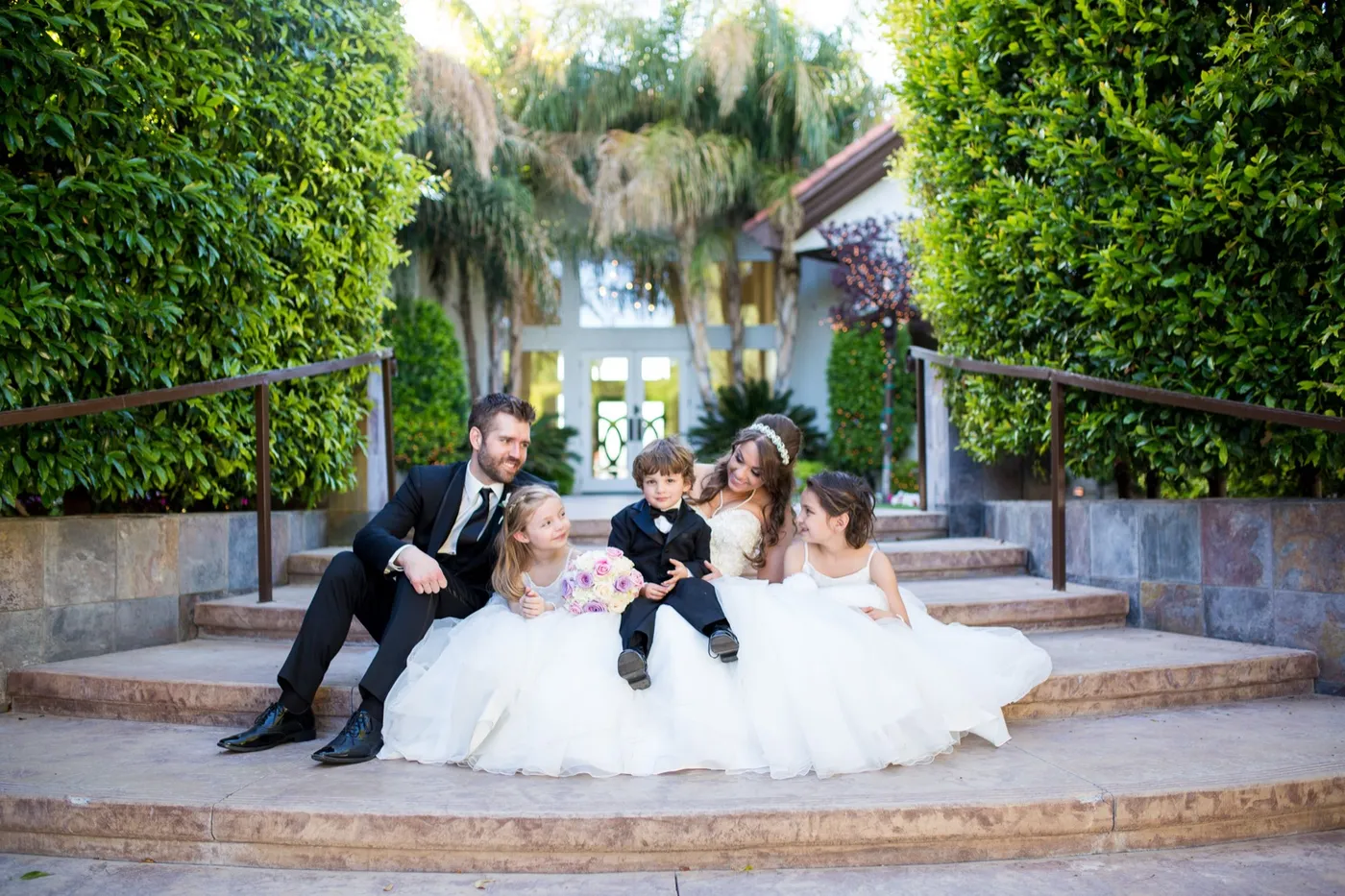 A bride and groom sit on wide garden steps between two flower girls in white and a small boy in a tuxedo