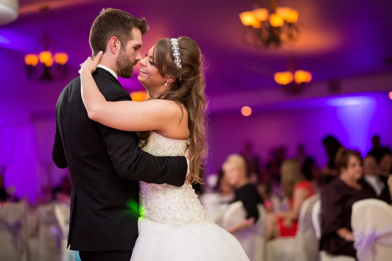 Couple dancing close together with guests seated around them and purple light across the room