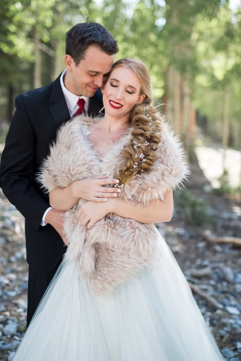 Groom holding his bride close as she wraps a fur stole around her shoulders in the trees