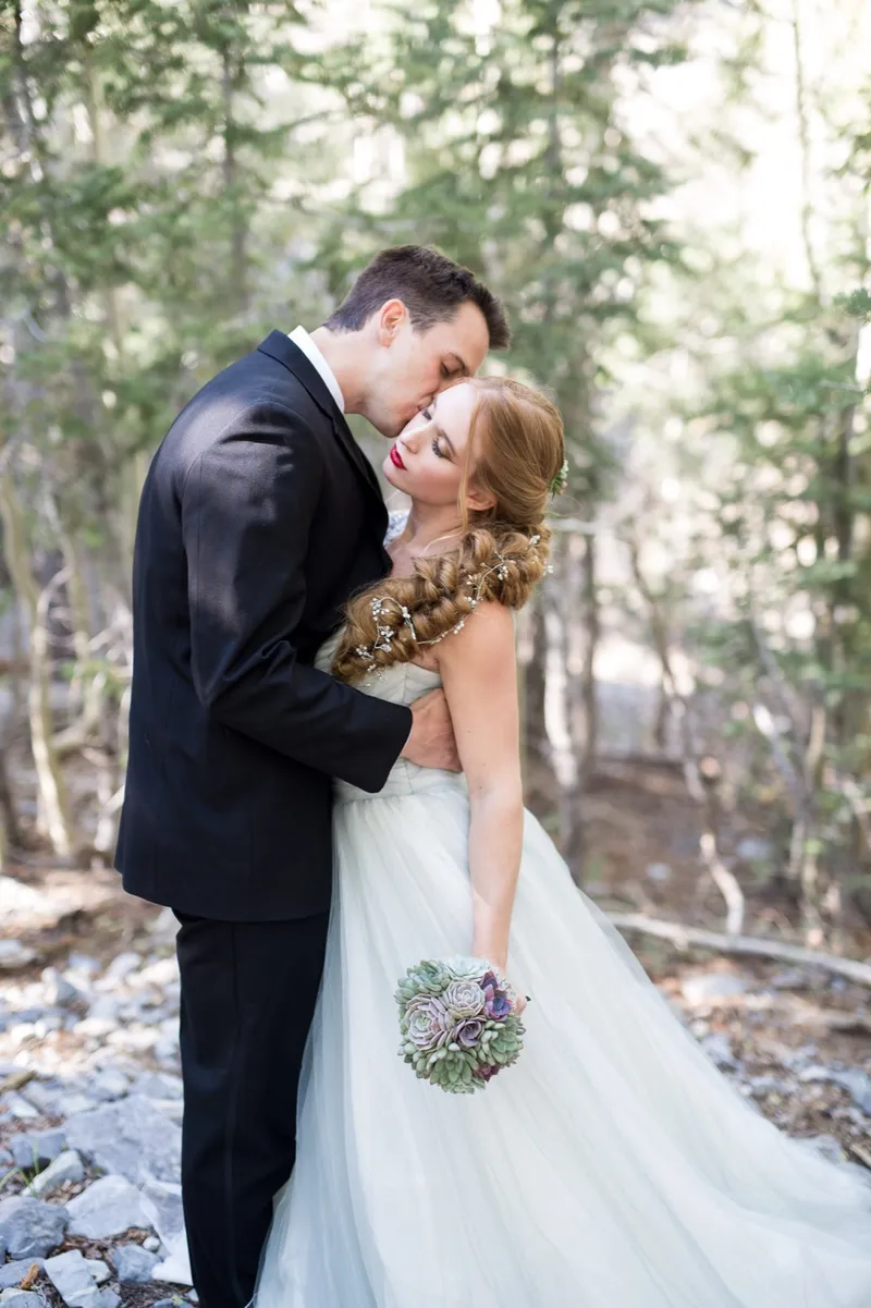 Groom kissing his bride's forehead in the pines with her succulent bouquet at her waist