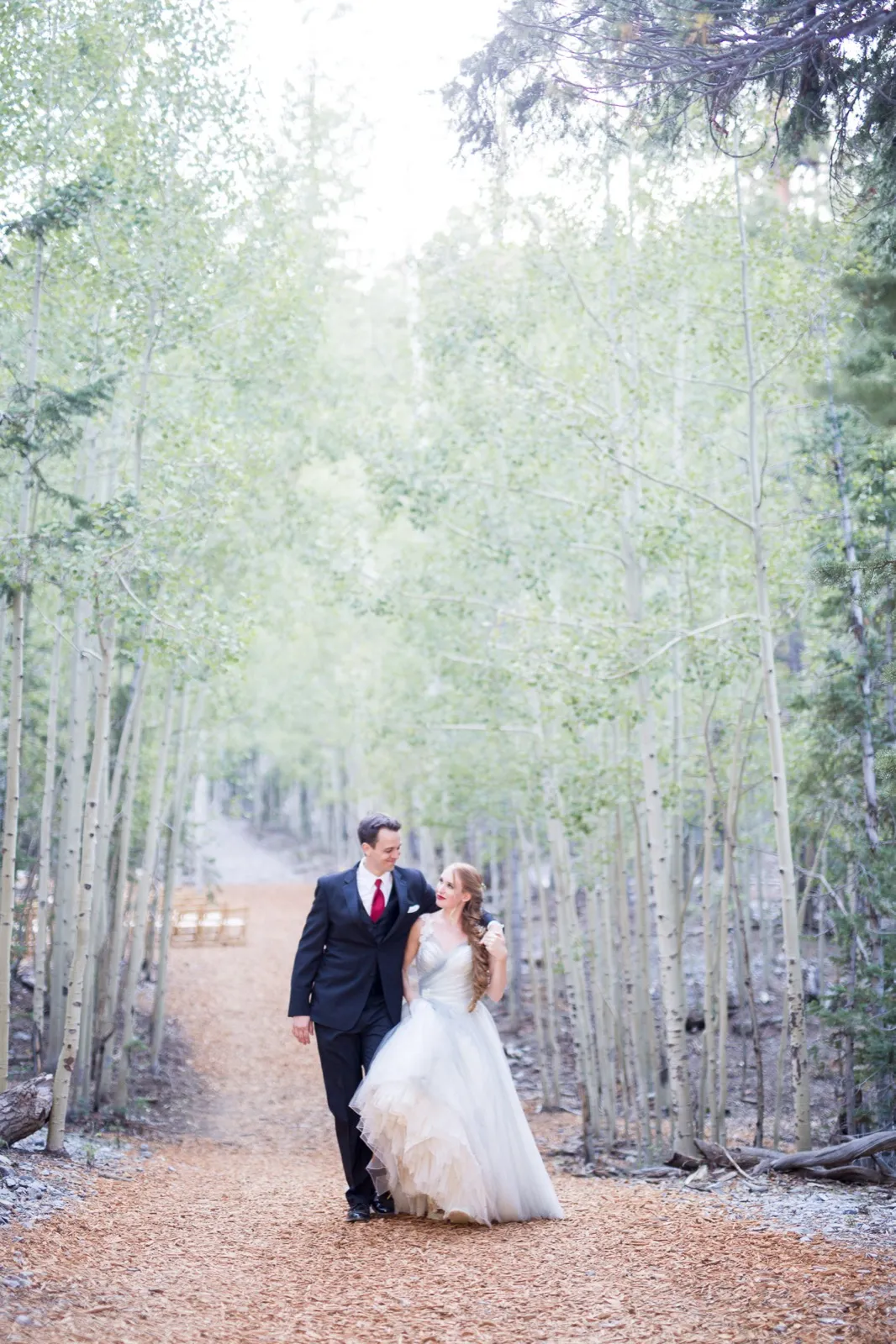 Bride and groom walk arm in arm down a pine needle path lined with aspens, gold ceremony chairs still set up behind them