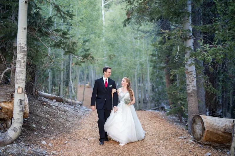 Bride and groom walking a wide forest road between white aspen trunks, looking at each other, cut logs at the edges