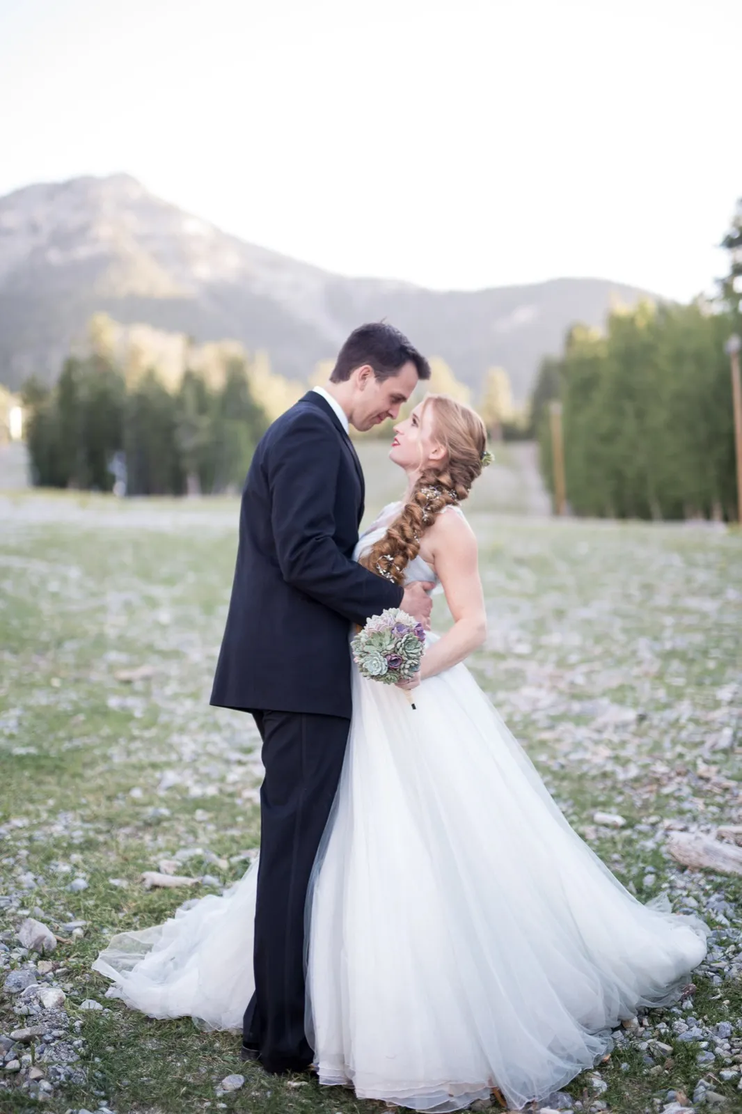 Bride and groom embracing in an open meadow as the last light catches the bare rock ridge above the pines