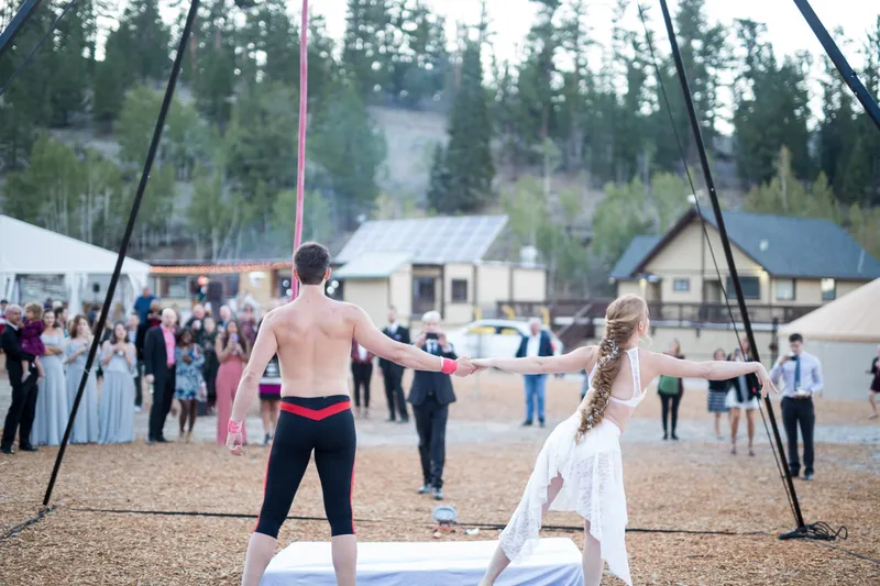 Two aerial performers standing hand in hand on a mat under their rig at dusk while wedding guests watch, a white tent and buildings behind them