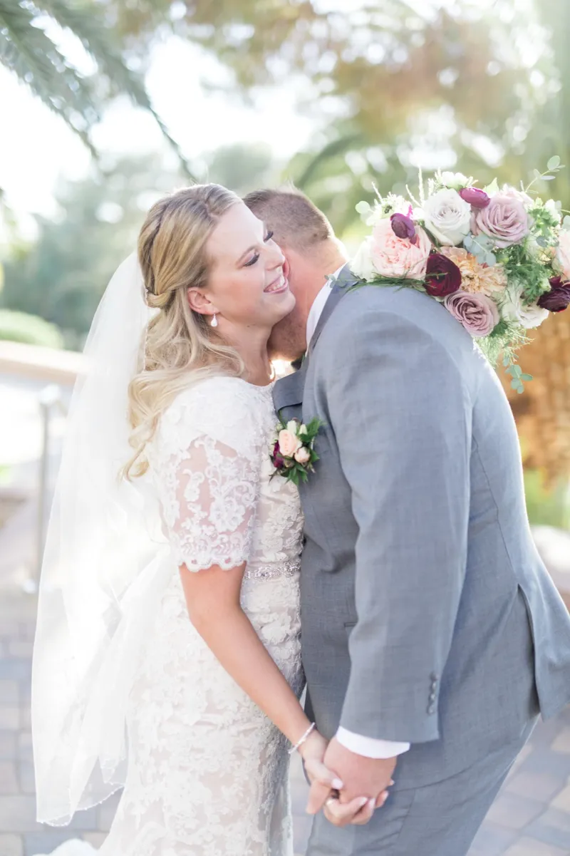 The groom kissing the bride's cheek in a close frame, her veil behind her and her burgundy and blush bouquet raised beside them