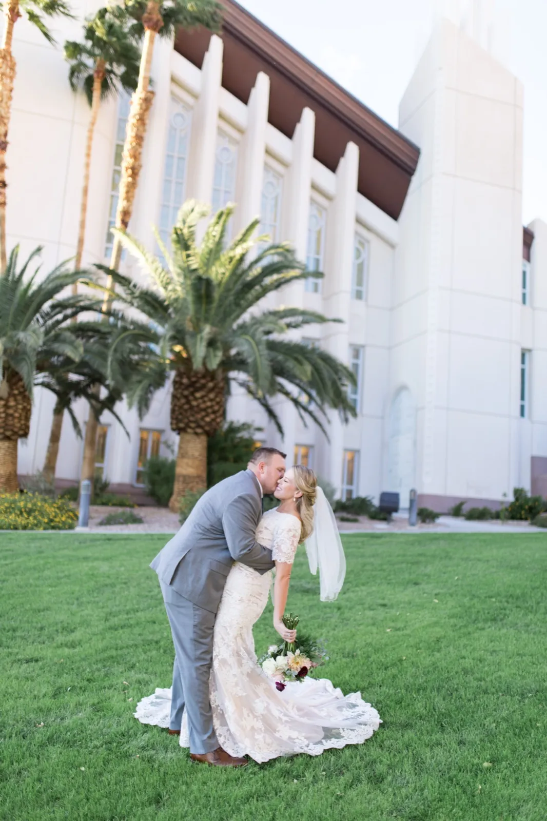 A groom dipping his bride for a kiss on the grass beside the temple’s tall windows and date palms