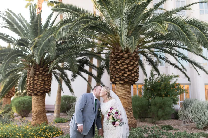 The couple kissing under a date palm with yellow flowering shrubs along the wall behind them