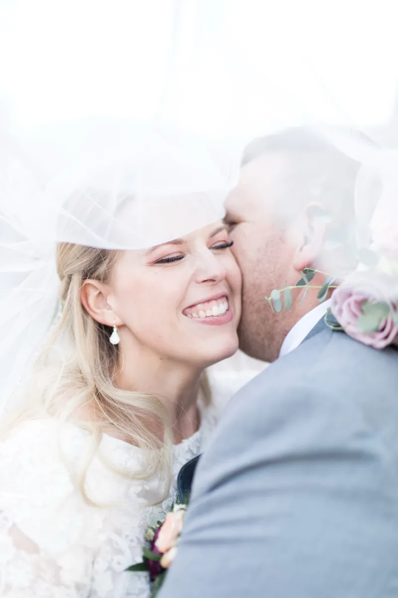 The bride laughing with her eyes closed as the groom kisses her cheek through her veil