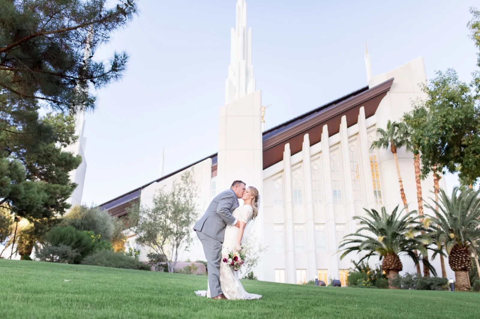 A couple kissing on the green lawn in front of the white temple, its tallest spire and gold statue above them and a row of date palms along the wall