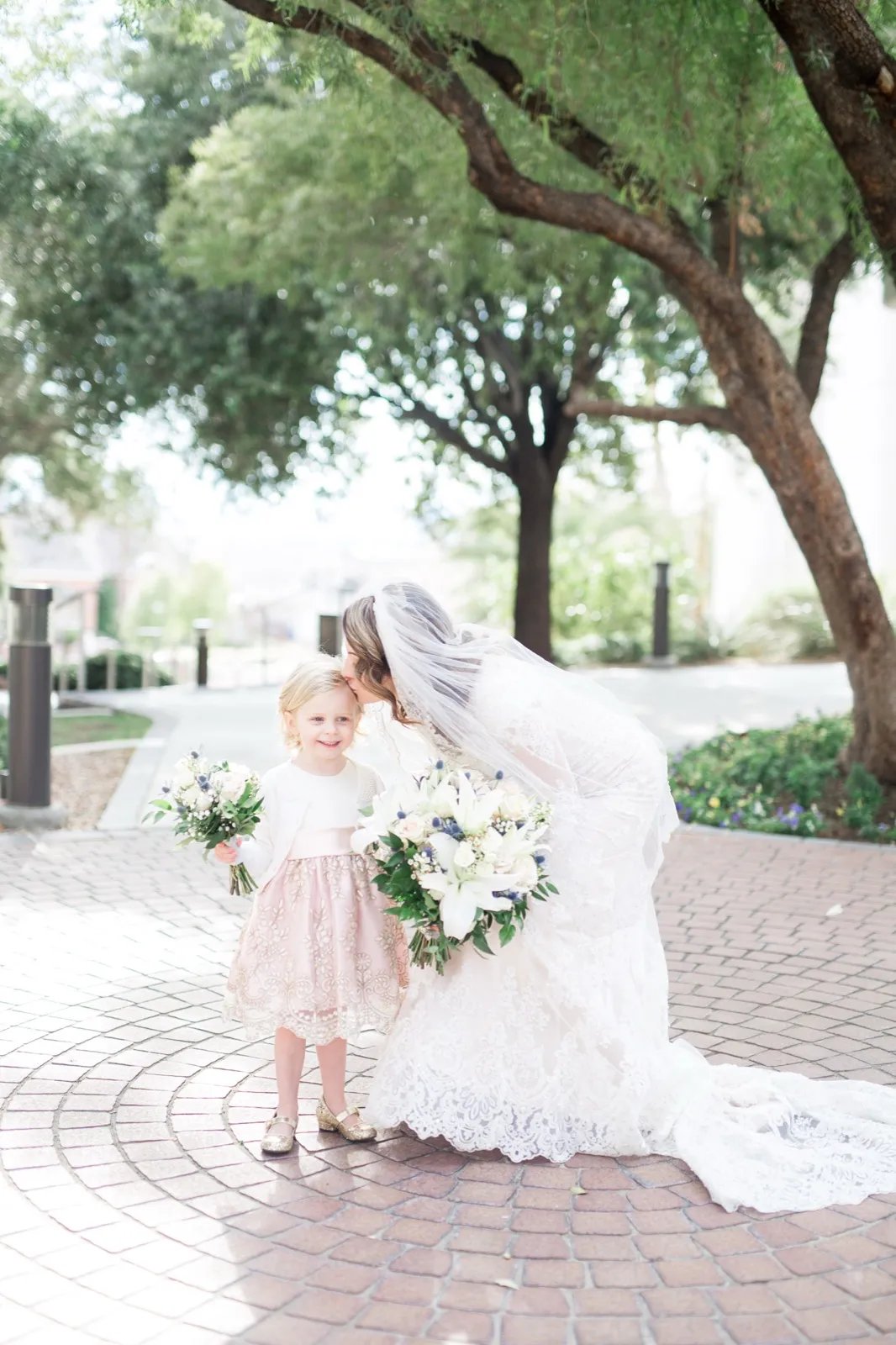 A bride bending down to kiss a small flower girl on the head, both holding white bouquets