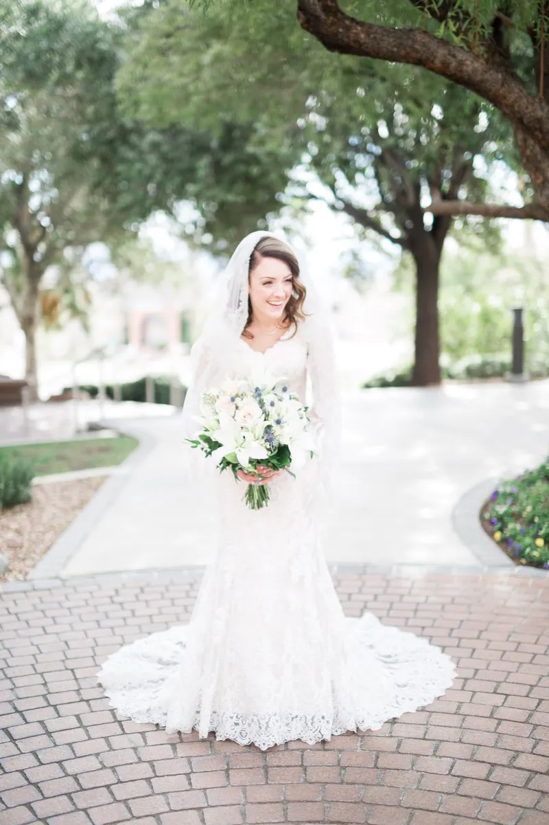 The bride alone on the round paver plaza under the trees with her train laid out and a white bouquet in her hands