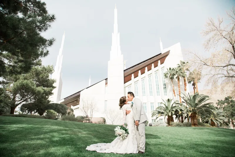 A wide frame of the couple standing on the lawn with the temple's white spires above the trees behind them