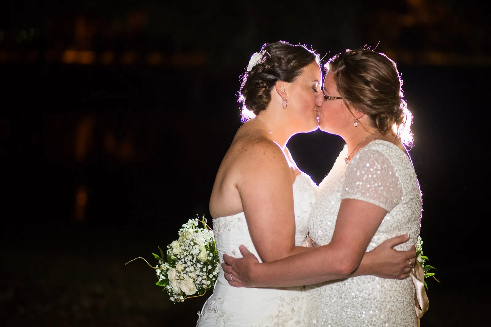 Two brides in beaded gowns kissing outdoors at night, rim lit against dark trees, one holding a white bouquet