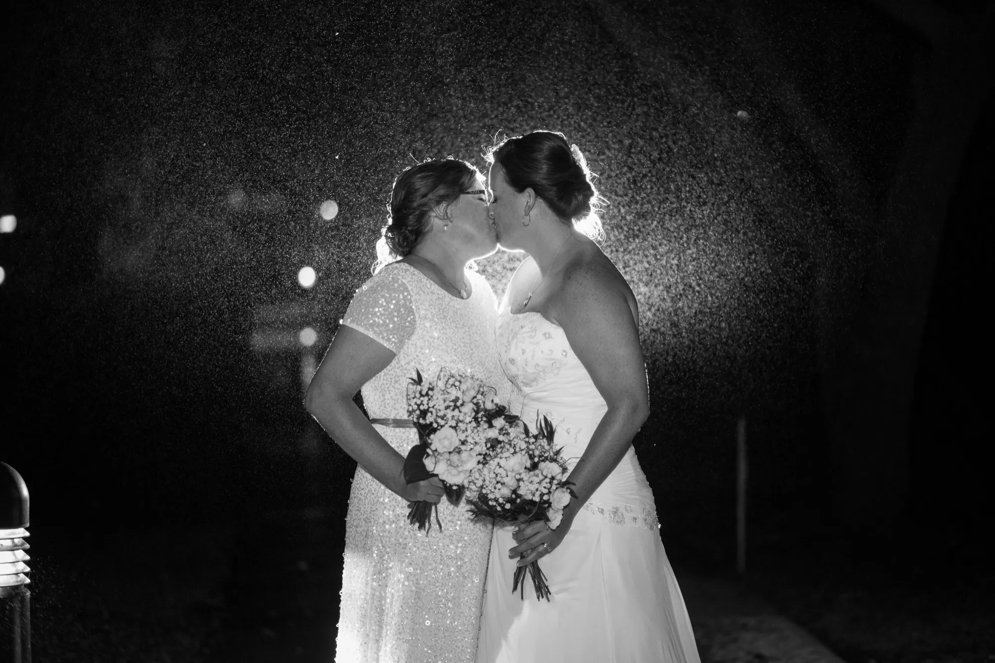 Two brides kissing in the rain at night, photographed in black and white