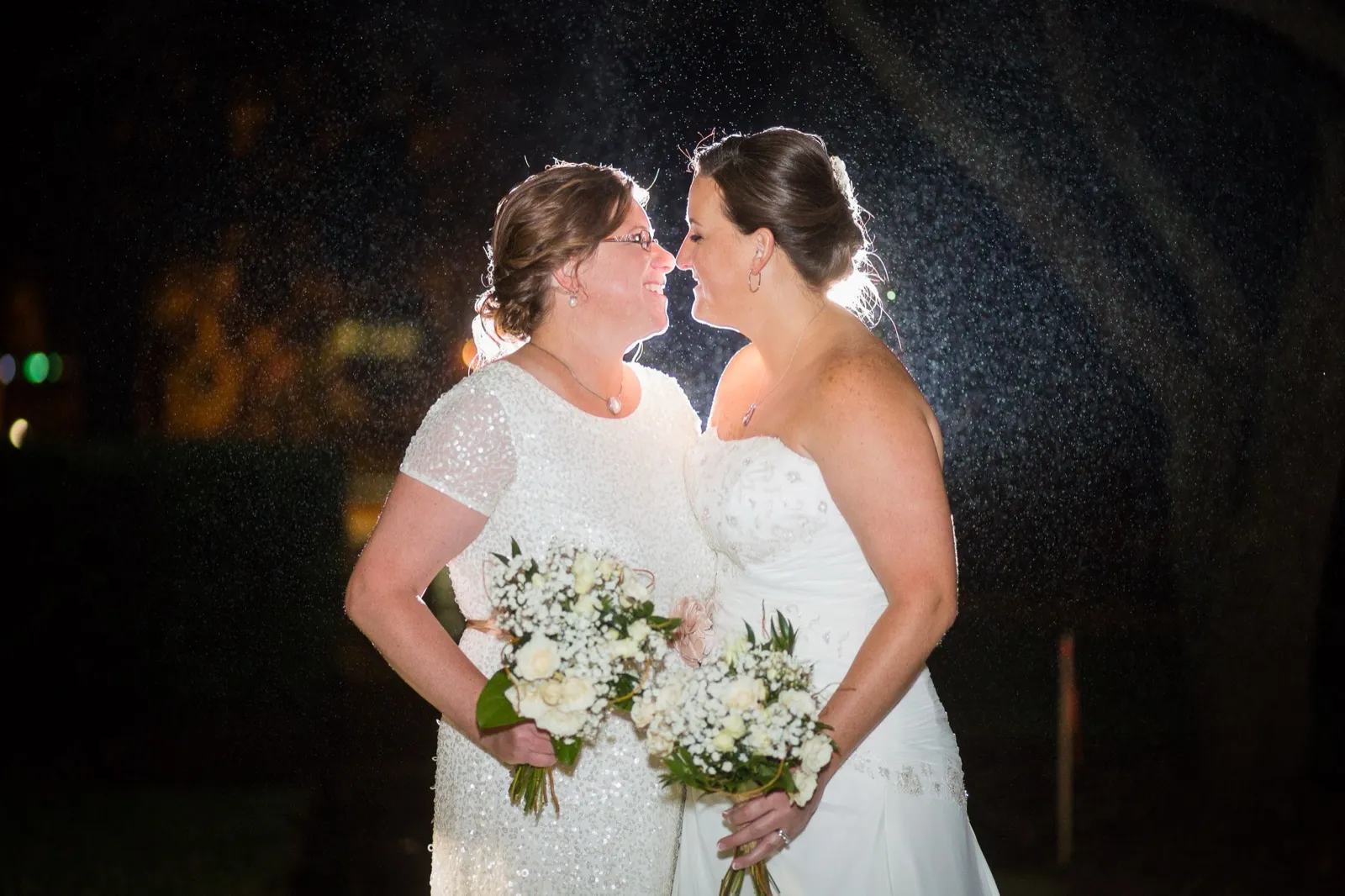 Two brides nose to nose at night holding white bouquets, only lamps and dark trees behind them