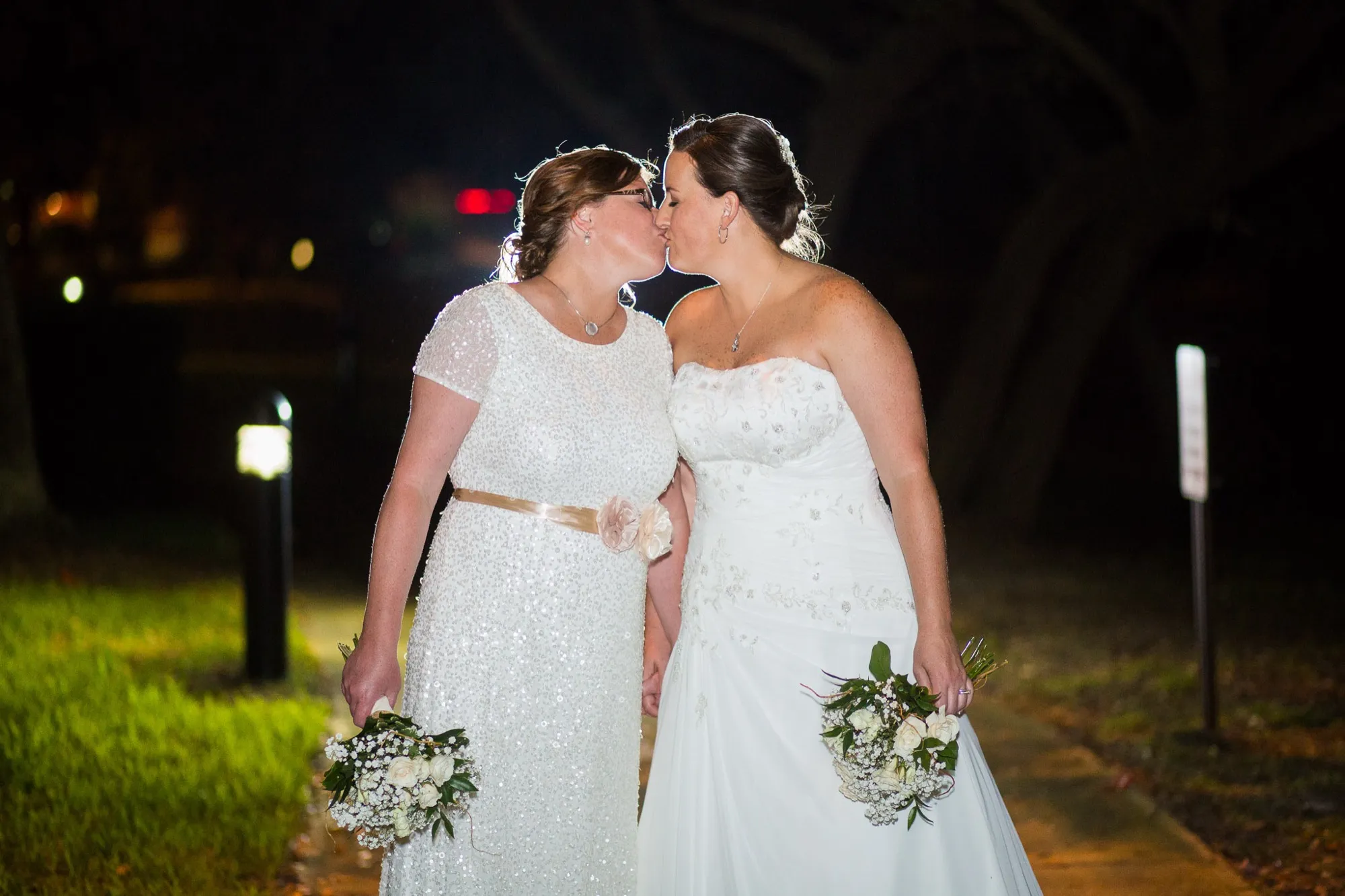 Two brides kissing at night with their bouquets, city lights glowing behind them