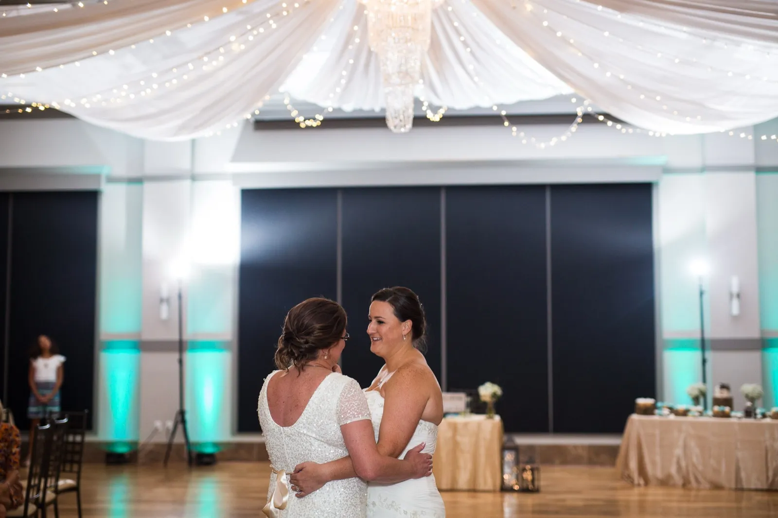 Two brides sharing their first dance in a ballroom under draped fabric and string lights