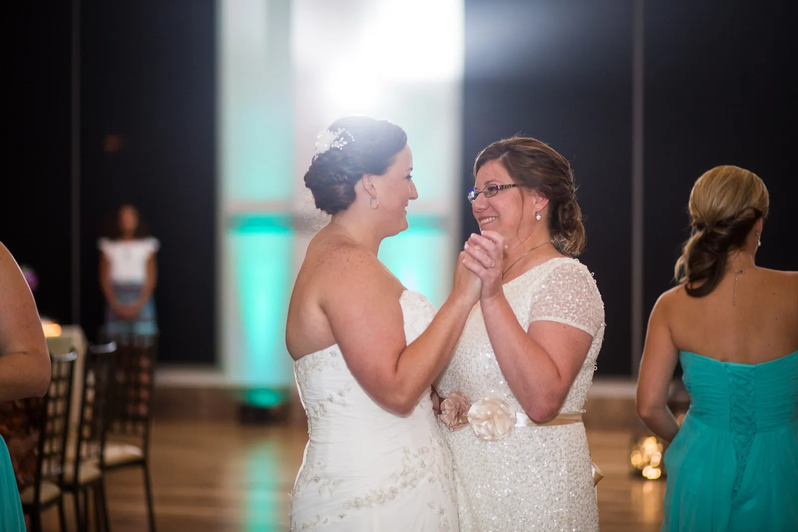 Two brides hold hands and smile at each other during their first dance on a ballroom floor lit teal