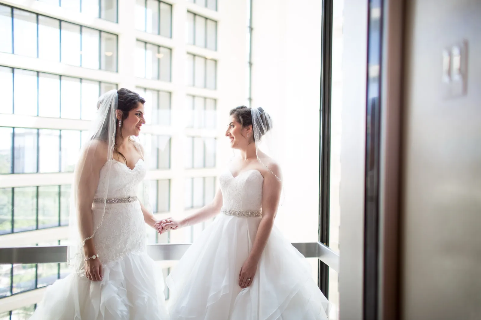 Two brides in gowns and veils hold hands and face each other beside tall windows before their ceremony