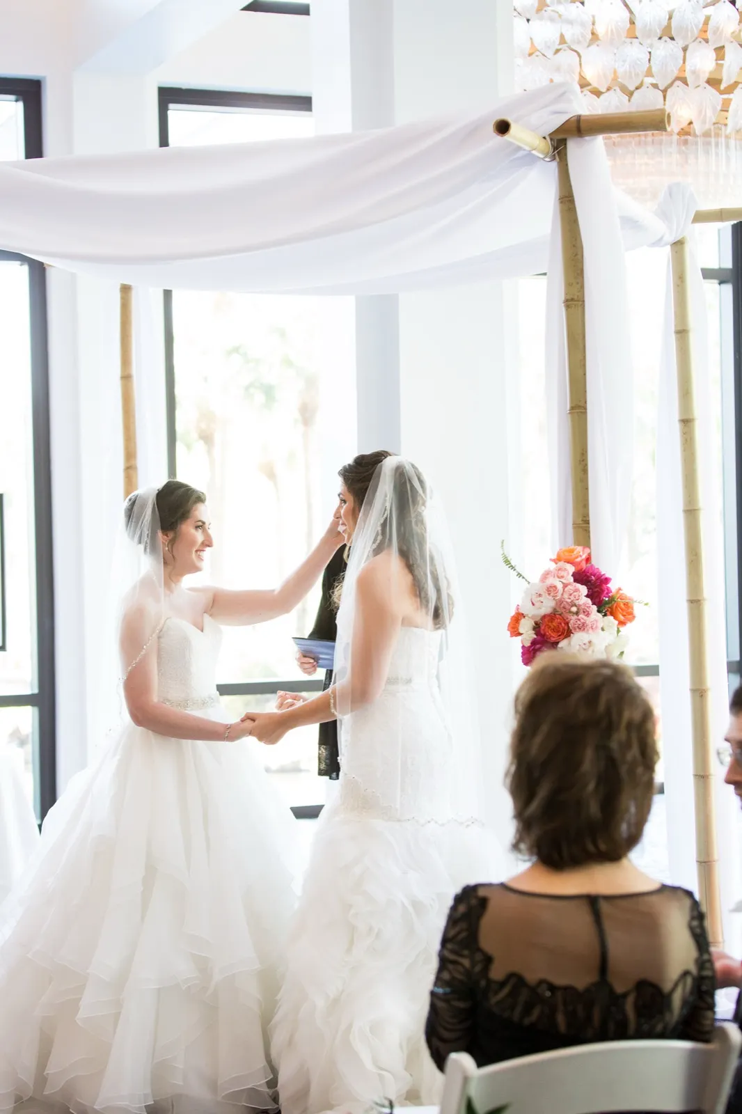 Two brides in white gowns holding hands under a draped bamboo chuppah, one reaching to touch the other’s veil