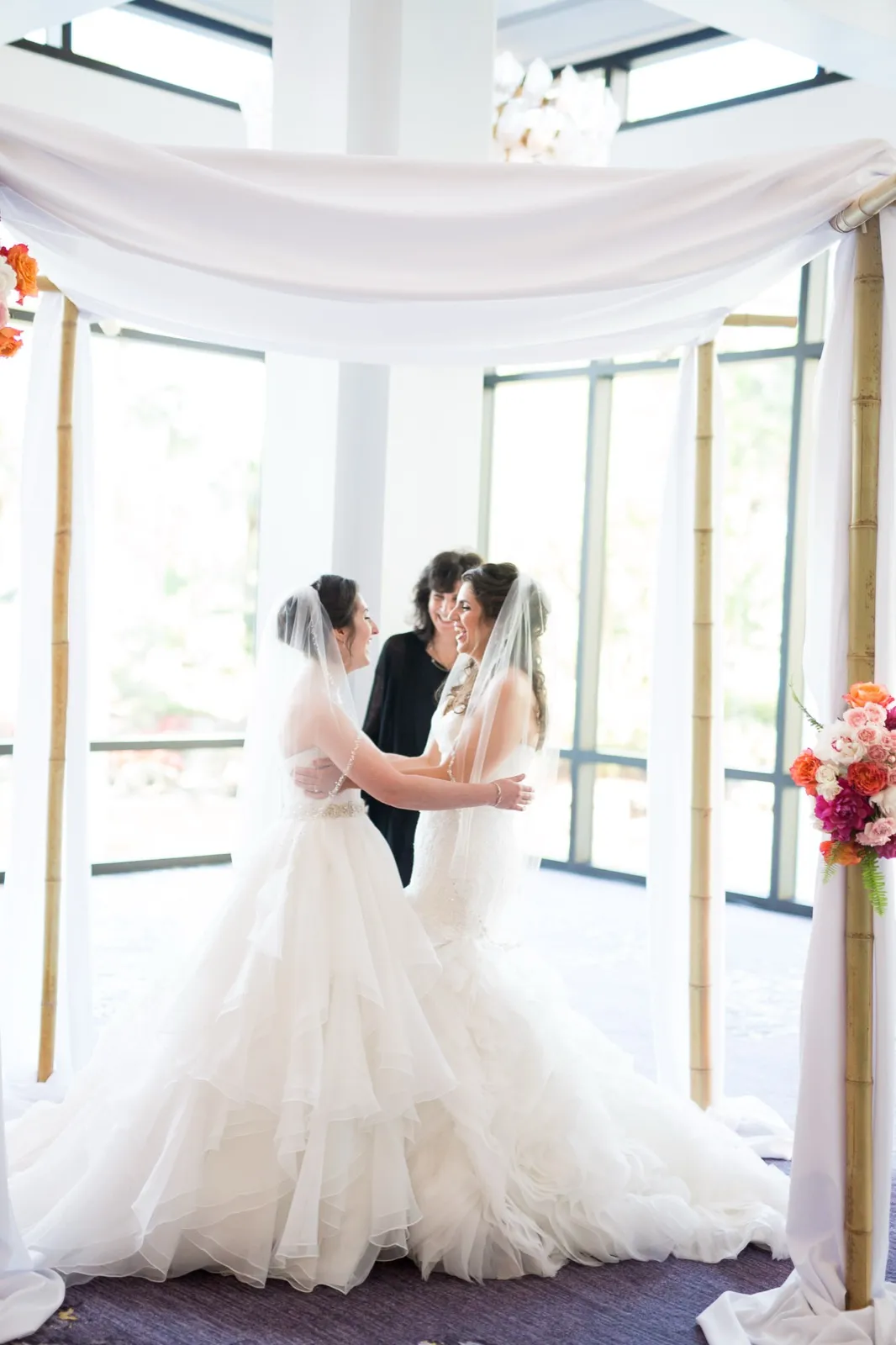 Two brides in white gowns and veils embrace and laugh under a draped bamboo chuppah while their officiant smiles behind them