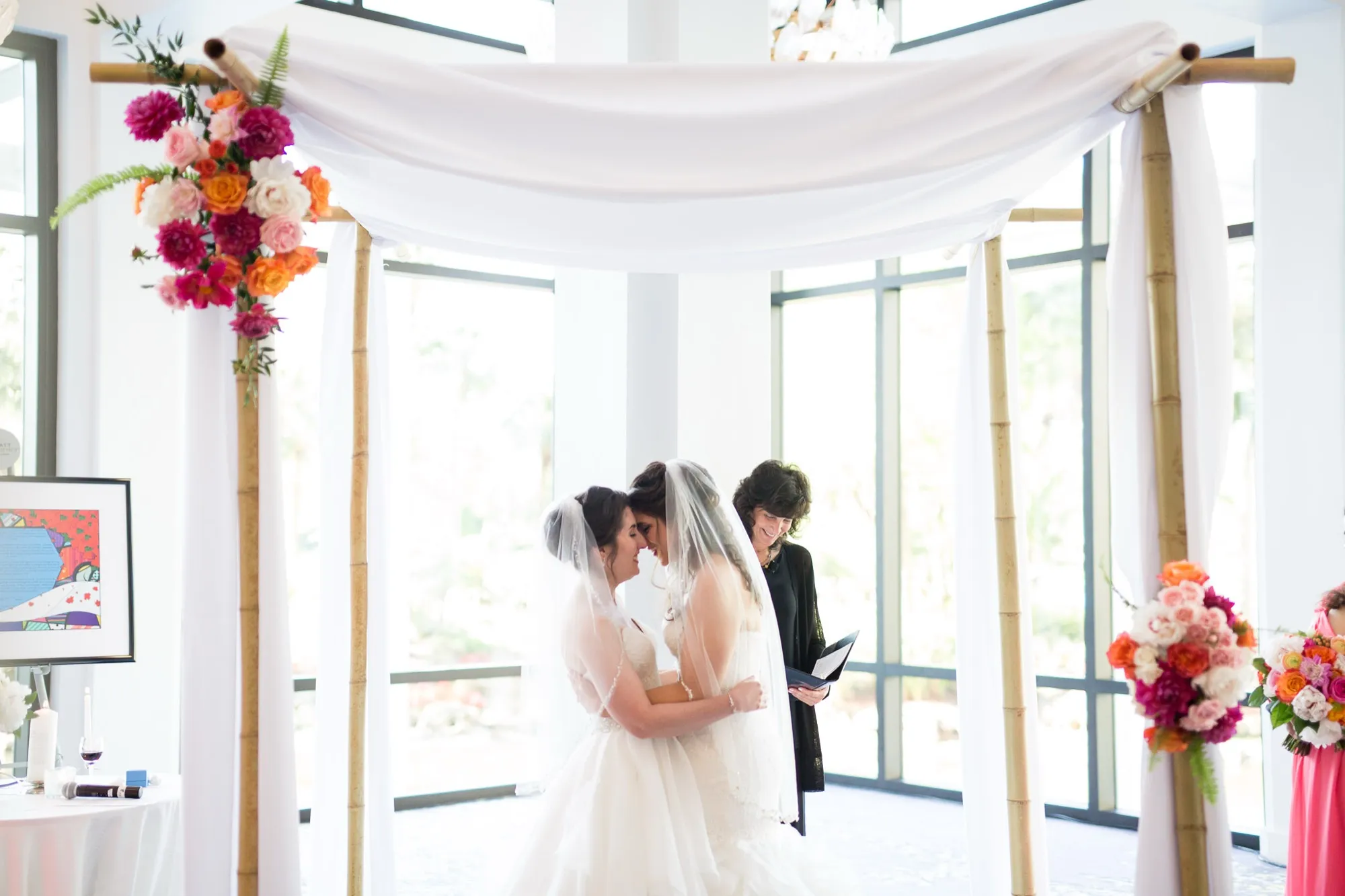 Two brides touching foreheads under a flower-draped canopy during their wedding ceremony