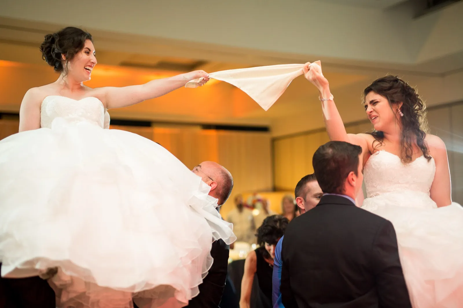 Two brides lifted on chairs during the hora, holding opposite ends of a napkin between them