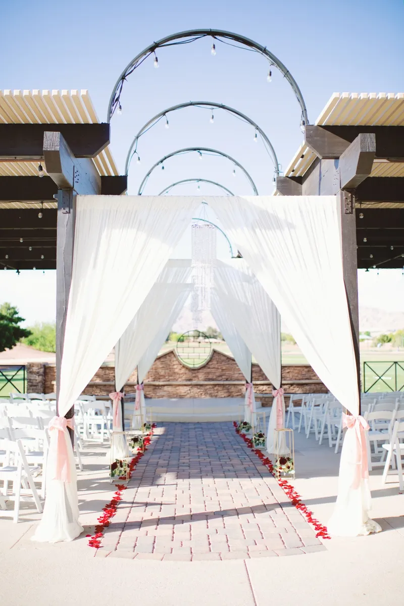 White draped arch under the covered patio at the end of a brick paver aisle scattered with red petals