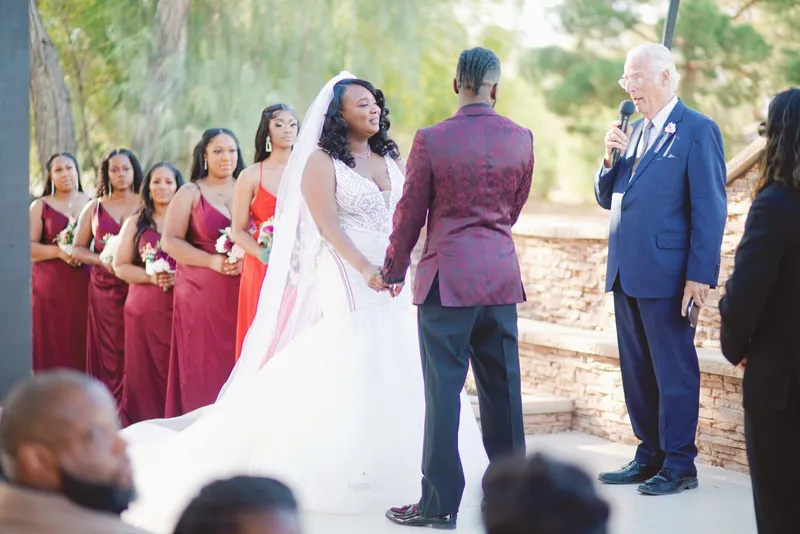 Couple holding hands at the ceremony with bridesmaids in burgundy and red lined up behind the bride and the officiant speaking