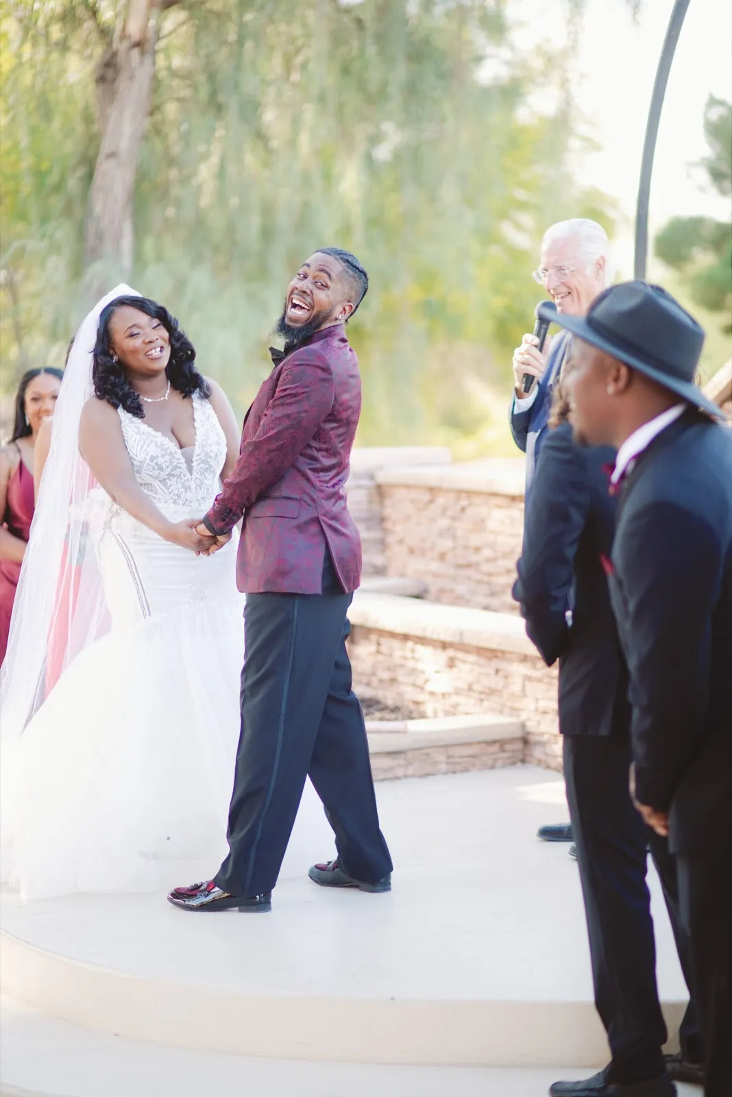 Black bride in a fitted white gown and groom in a burgundy tuxedo laughing on the ceremony platform as their officiant speaks into a microphone