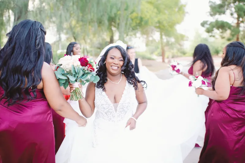 Bridesmaids in burgundy crouching to fan out the bride's gown while she laughs at the camera