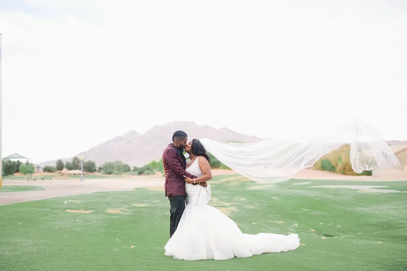 Bride and groom kiss on the golf course as her long veil streams sideways, mountains behind them