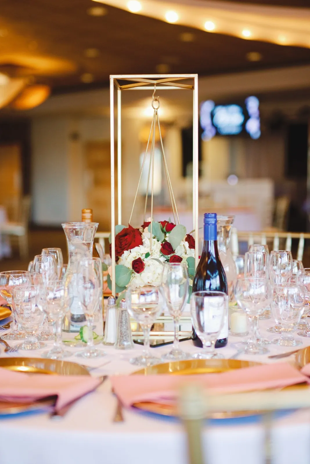 Tall gold frame centerpiece of red roses and white hydrangea above gold chargers and blush napkins