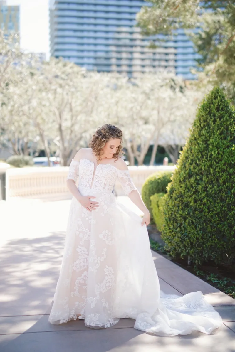 Bride in an off the shoulder lace gown gathering her train on the sidewalk beside the hedges