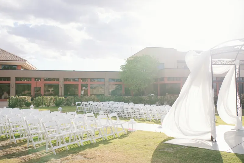 Rows of white chairs set on the grass facing a white draped arch, one panel of the drape lifting in the wind and the clubhouse behind