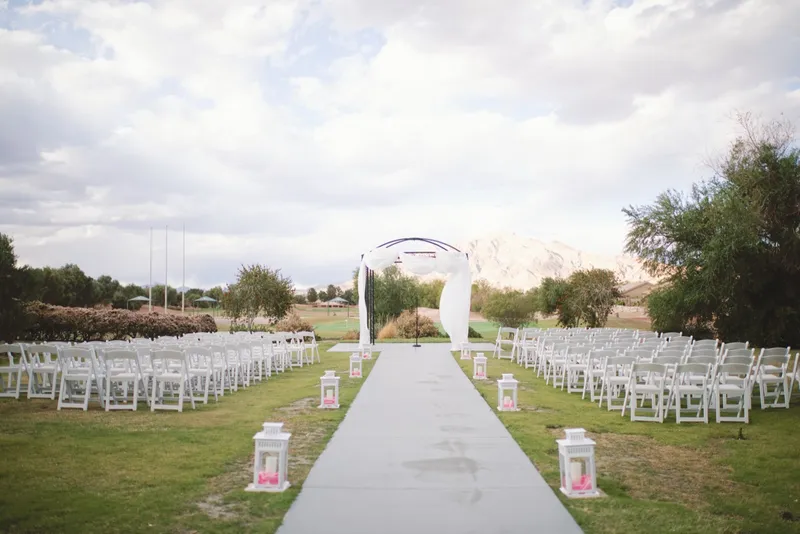 Lawn ceremony set with white chairs and a paved aisle lined with lanterns leading to a white draped arch, mountains in the distance