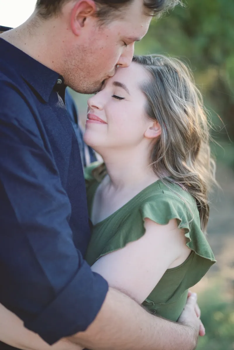 Man kissing his fiancee on the forehead with her eyes closed and green brush filling the background