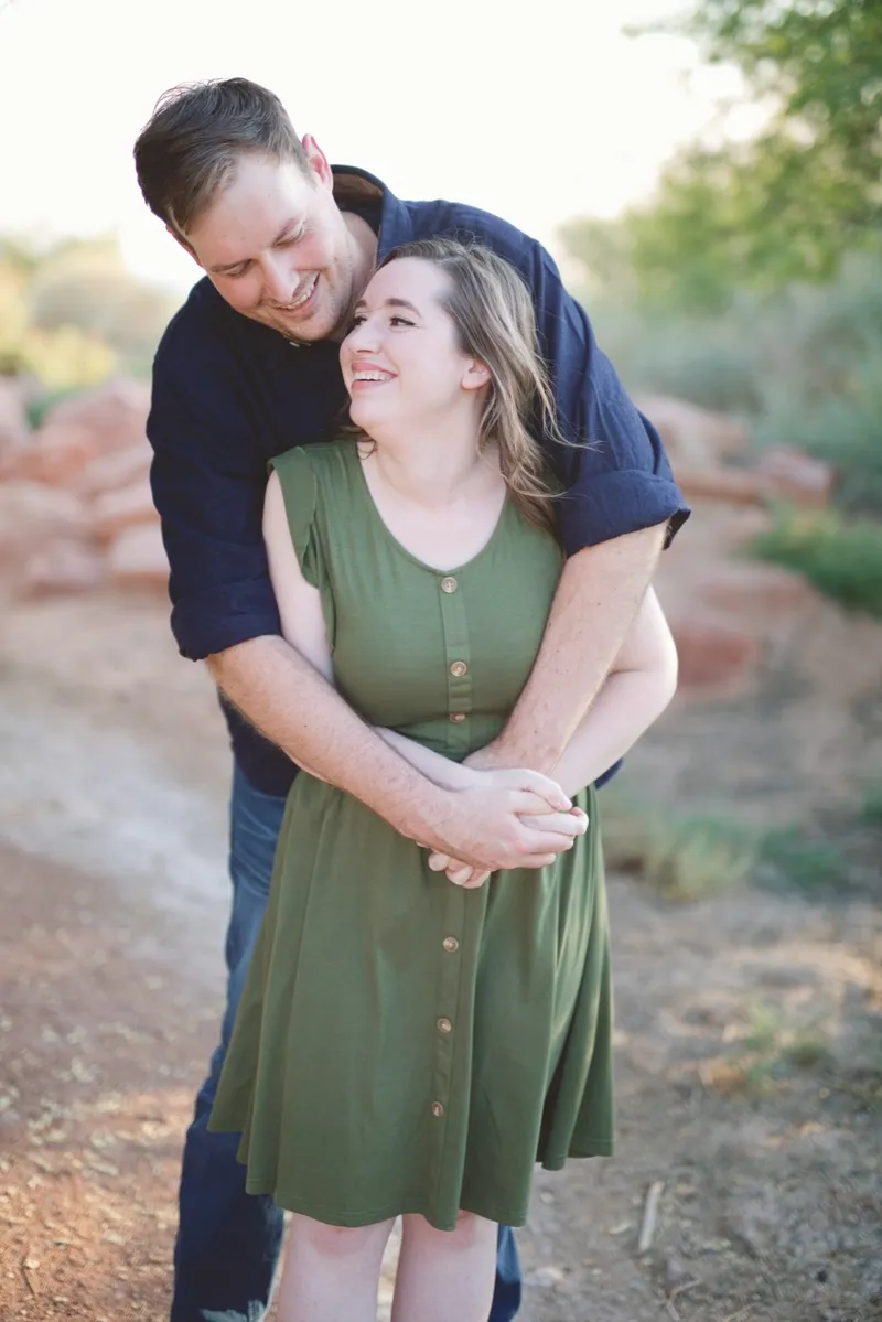 Man leaning over his fiancee's shoulder from behind on the trail while she laughs, with red landscaping rock along the edge of the path